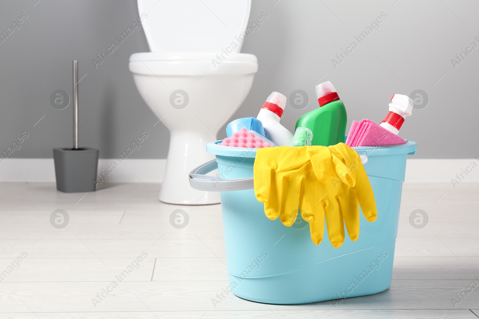 Photo of Bucket with different toilet cleaners, rag, sponge and gloves on floor in bathroom