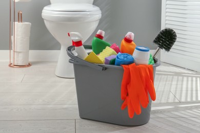 Photo of Bucket with different toilet cleaners, sponges, brush and gloves on floor in bathroom