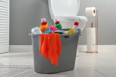 Photo of Bucket with different toilet cleaners, sponge and gloves on floor in bathroom