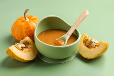 Photo of Delicious baby food with spoon in bowl and fresh pumpkins on green table