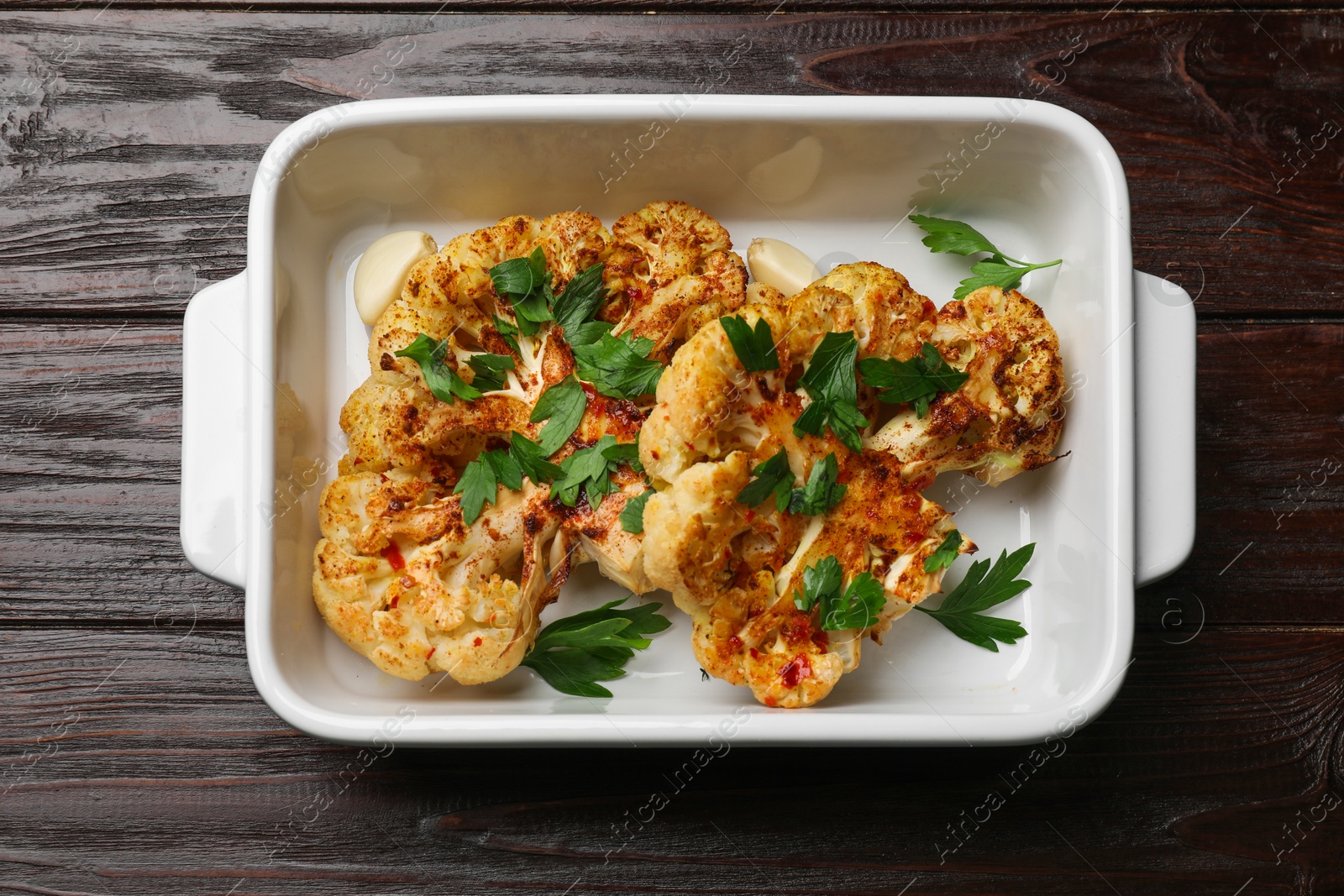 Photo of Tasty cauliflower steaks and parsley in baking dish on wooden table, top view