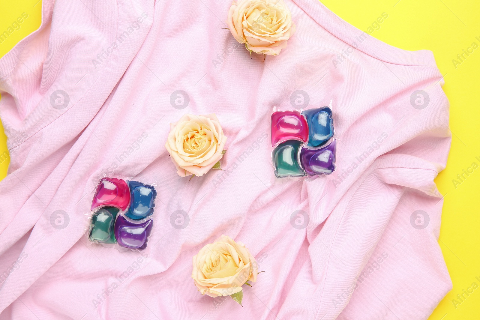 Photo of Laundry detergent pods, t-shirt and flowers on yellow background, flat lay