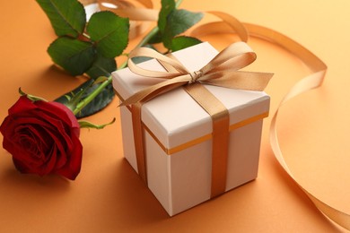 Photo of Beautiful gift box with golden bow and red rose on orange background, closeup