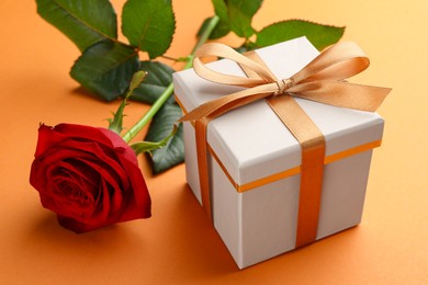 Photo of Beautiful gift box with golden bow and red rose on orange background, closeup