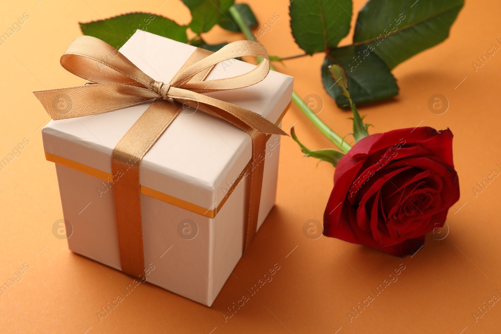 Photo of Beautiful gift box with golden bow and red rose on orange background, closeup