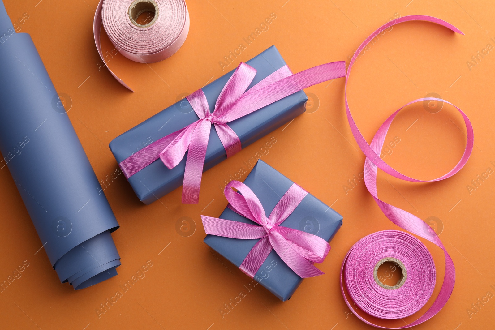 Photo of Beautiful gift boxes with pink bows, ribbons and roll of wrapping paper on orange background, flat lay