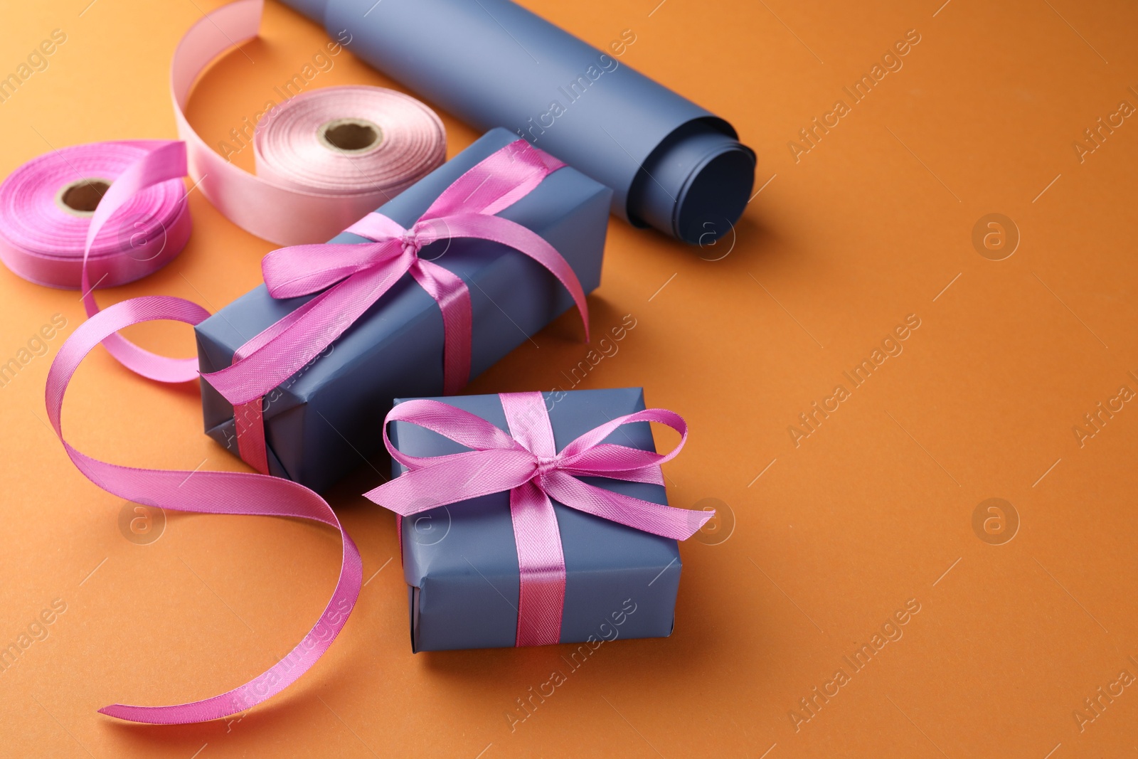 Photo of Beautiful gift boxes with pink bows, ribbons and roll of wrapping paper on orange background, space for text