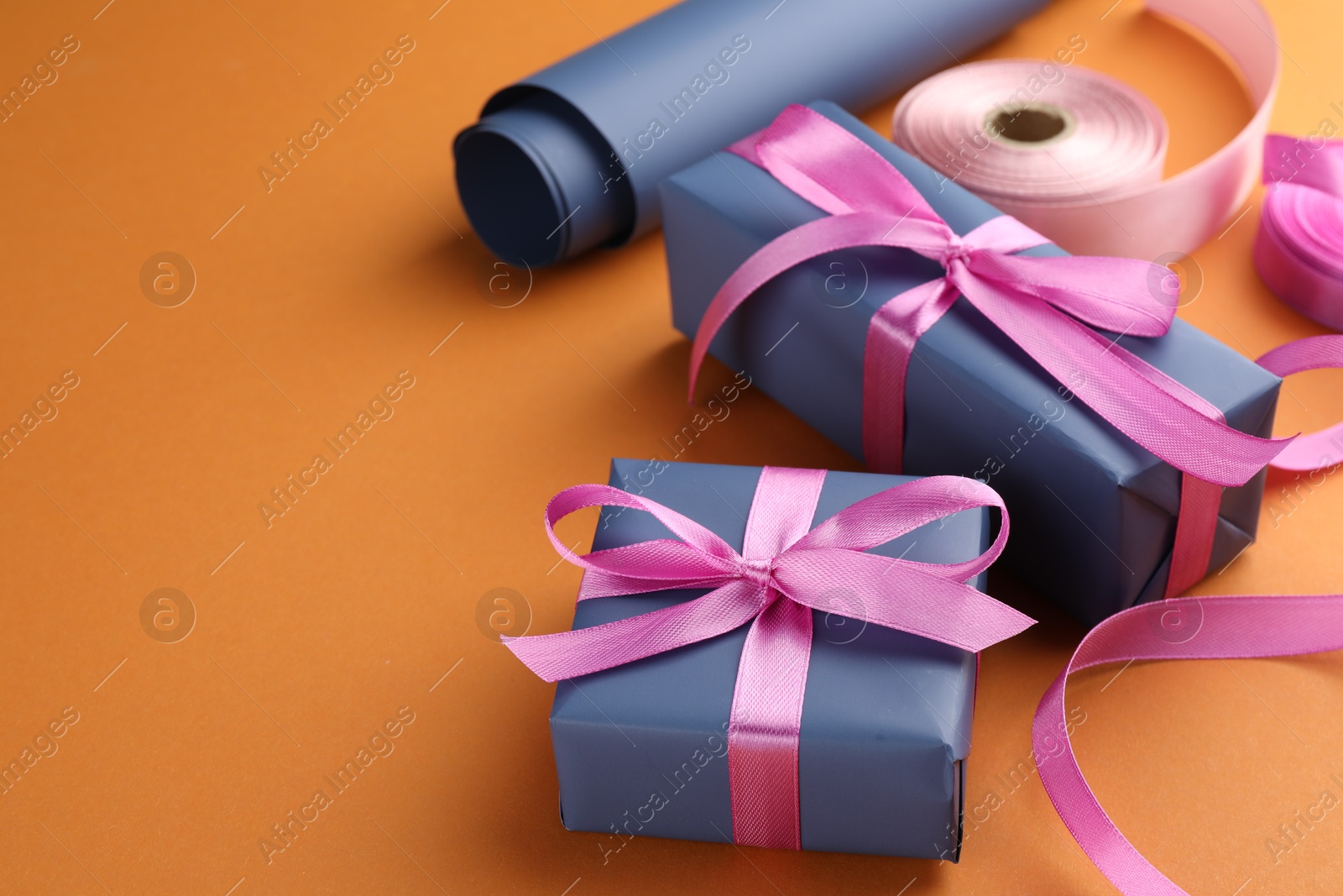 Photo of Beautiful gift boxes with pink bows, ribbons and roll of wrapping paper on orange background, space for text