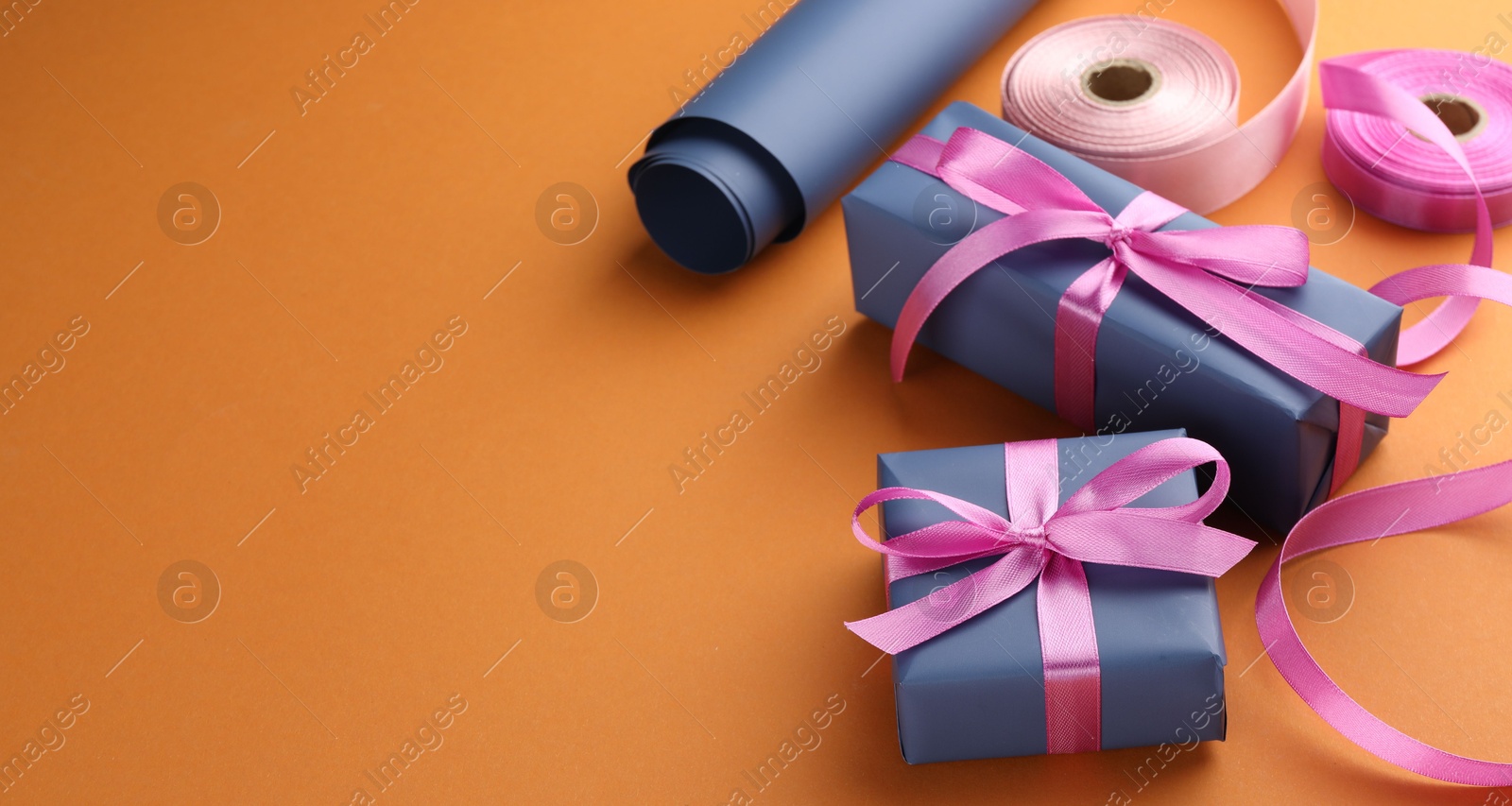 Photo of Beautiful gift boxes with pink bows, ribbons and roll of wrapping paper on orange background, space for text