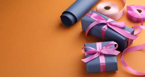 Beautiful gift boxes with pink bows, ribbons and roll of wrapping paper on orange background, space for text