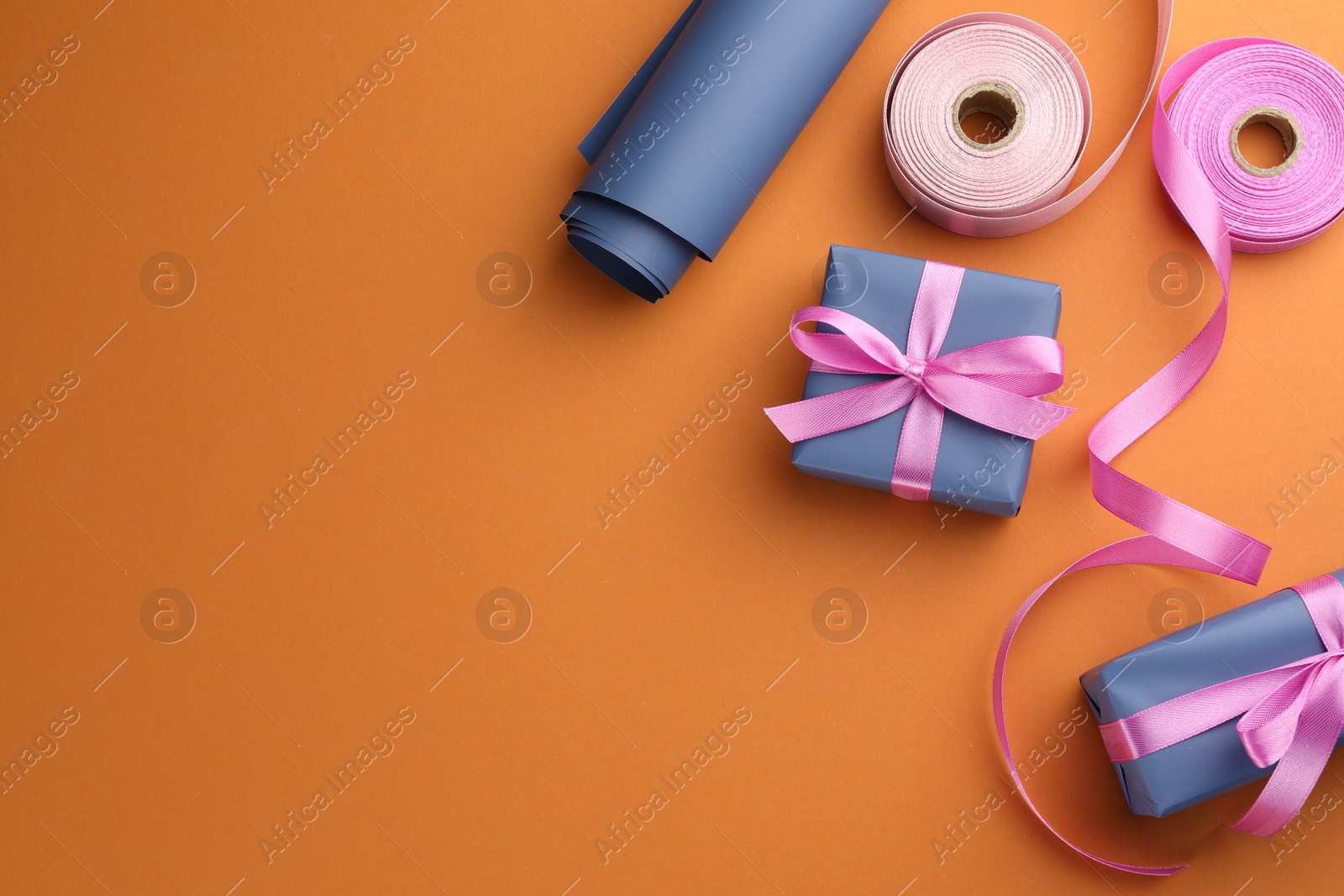 Photo of Beautiful gift boxes with pink bows, ribbons and roll of wrapping paper on orange background, flat lay. Space for text