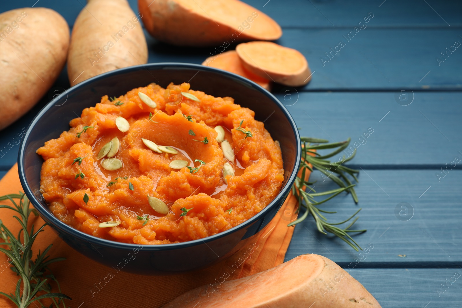 Photo of Tasty mashed sweet potato with pumpkin seeds in bowl, fresh vegetables and rosemary on blue wooden table, closeup