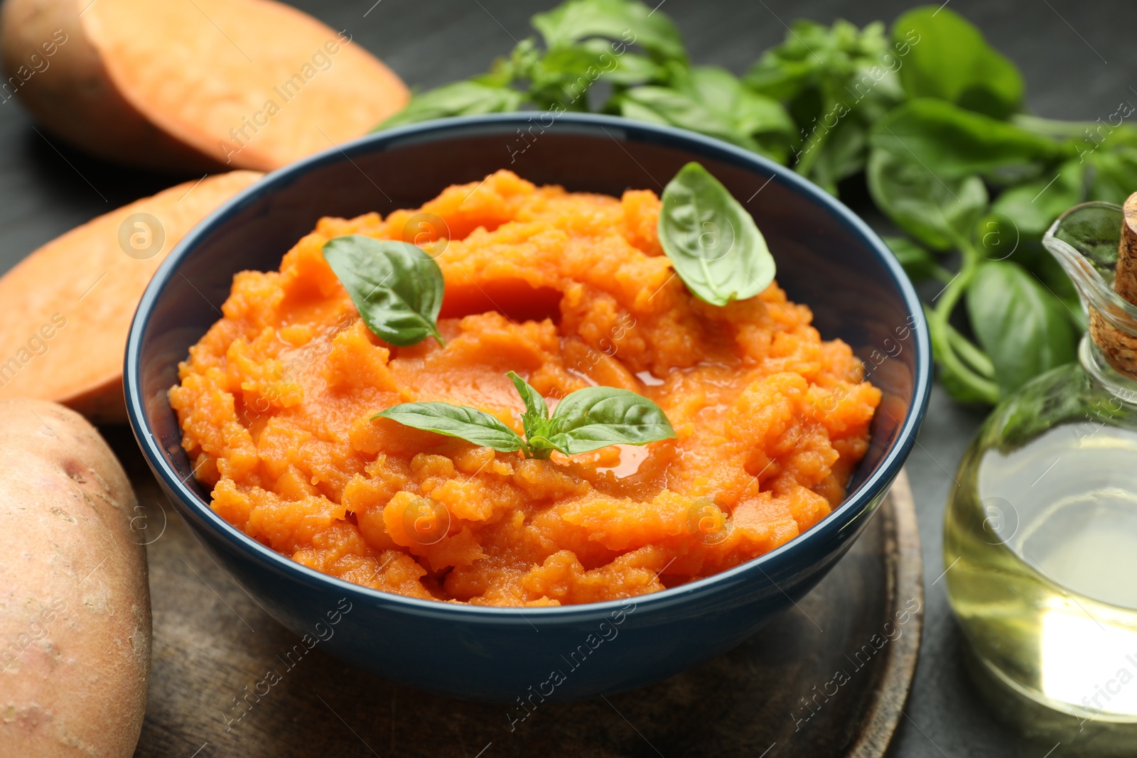 Photo of Tasty mashed sweet potato with basil in bowl, fresh vegetables and oil on dark table, closeup