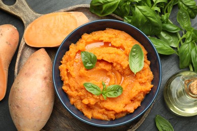 Photo of Tasty mashed sweet potato with basil in bowl, fresh vegetables and oil on dark gray table, flat lay