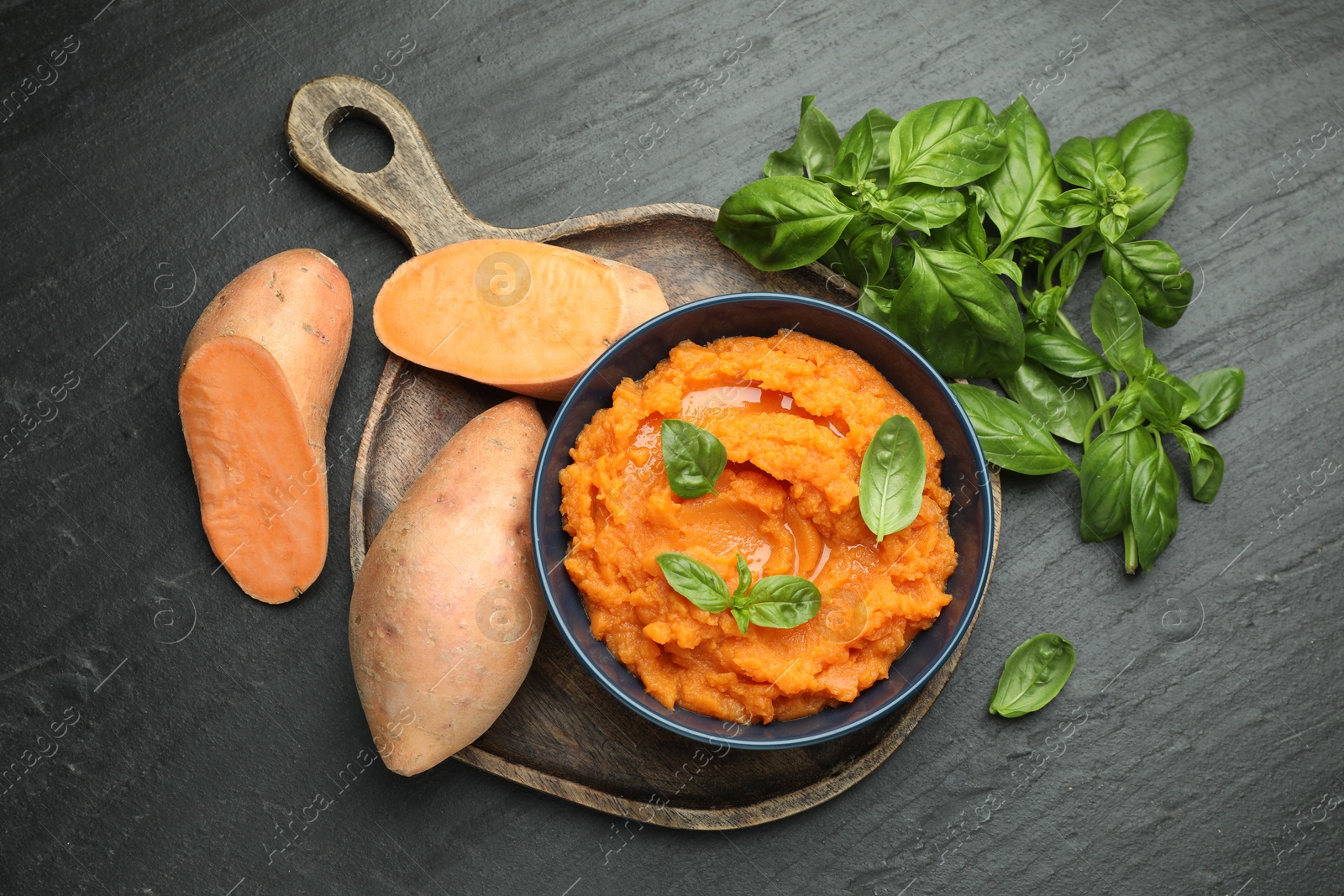 Photo of Tasty mashed sweet potato with basil in bowl and fresh vegetables on dark gray textured table, flat lay