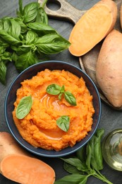 Photo of Tasty mashed sweet potato with basil in bowl and fresh vegetables on dark gray textured table, flat lay