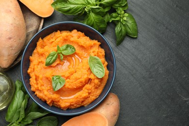 Photo of Tasty mashed sweet potato with basil in bowl and fresh vegetables on dark gray textured table, flat lay. Space for text