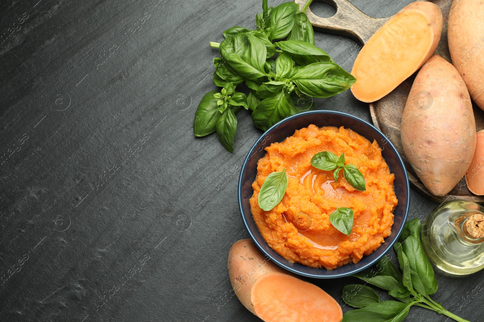 Photo of Tasty mashed sweet potato with basil in bowl, fresh vegetables and oil on dark gray textured table, flat lay. Space for text