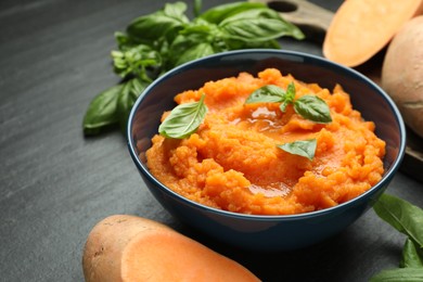 Photo of Tasty mashed sweet potato with basil in bowl and fresh vegetables on dark gray textured table, closeup