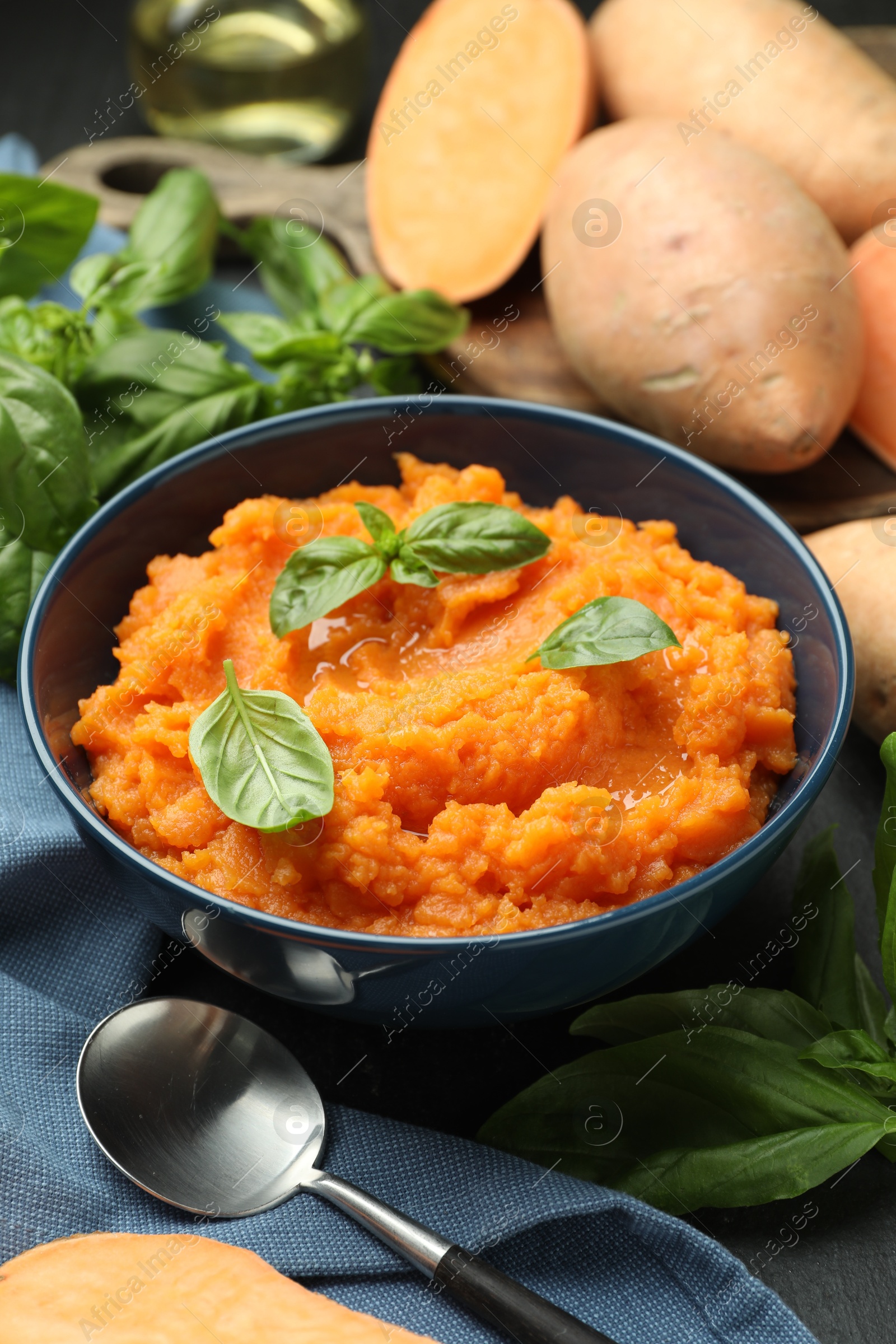 Photo of Tasty mashed sweet potato with basil in bowl, fresh vegetables and spoon on table, closeup