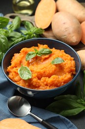 Photo of Tasty mashed sweet potato with basil in bowl, fresh vegetables and spoon on table, closeup