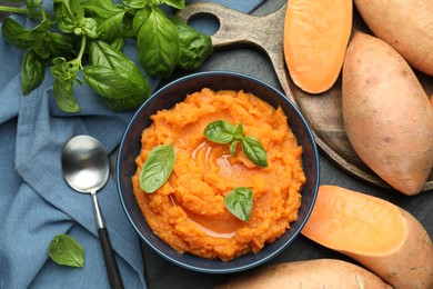 Photo of Tasty mashed sweet potato with basil in bowl, fresh vegetables and spoon on table, flat lay