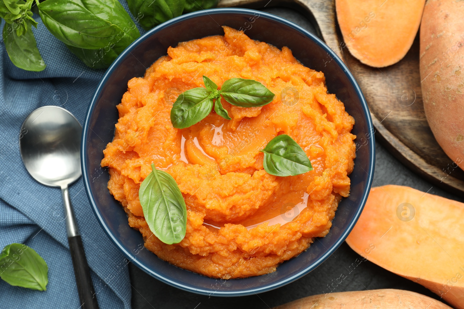 Photo of Tasty mashed sweet potato with basil in bowl, fresh vegetables and spoon on table, flat lay