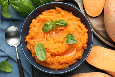 Photo of Tasty mashed sweet potato with basil in bowl, fresh vegetables and spoon on table, flat lay