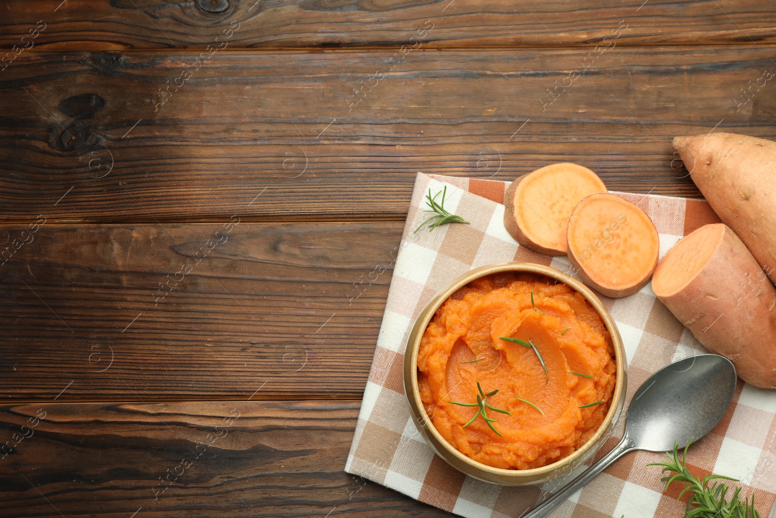 Photo of Tasty mashed sweet potato with rosemary in bowl, fresh vegetables and spoon on wooden table, flat lay. Space for text