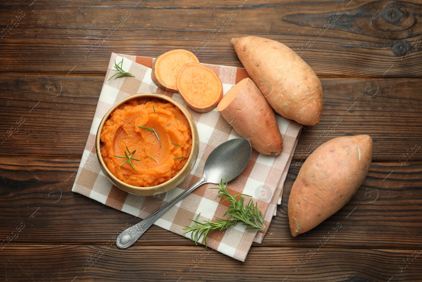 Photo of Tasty mashed sweet potato with rosemary in bowl, fresh vegetables and spoon on wooden table, flat lay