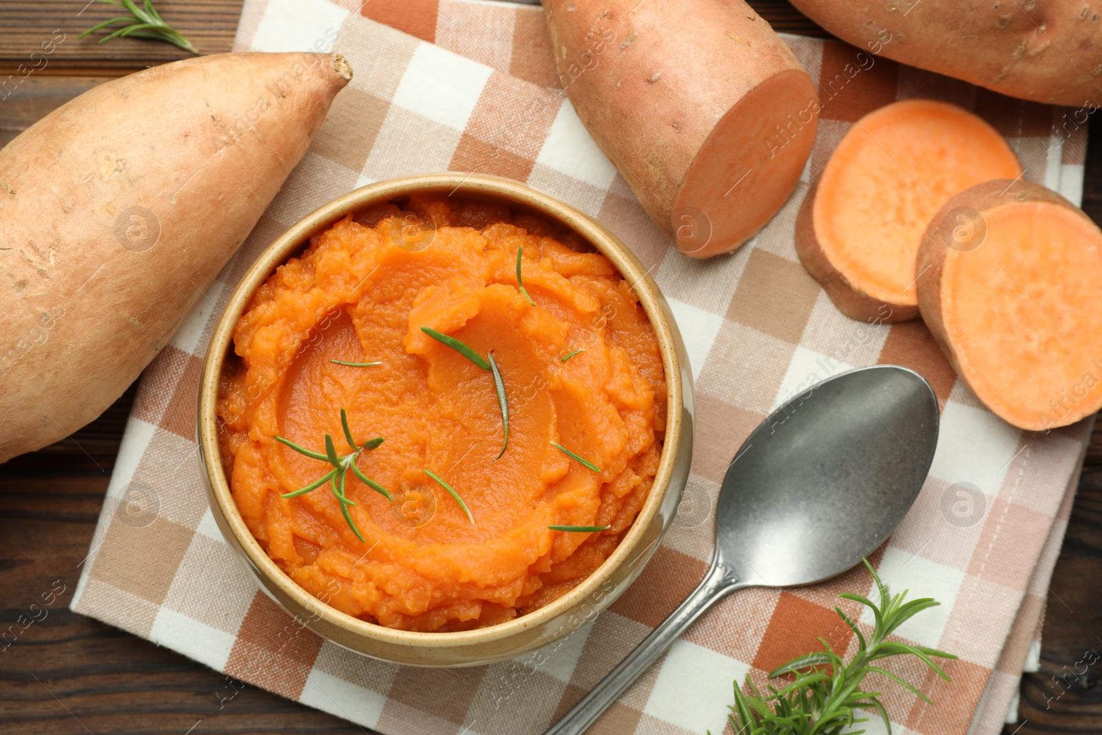 Photo of Tasty mashed sweet potato with rosemary in bowl, fresh vegetables and spoon on wooden table, flat lay