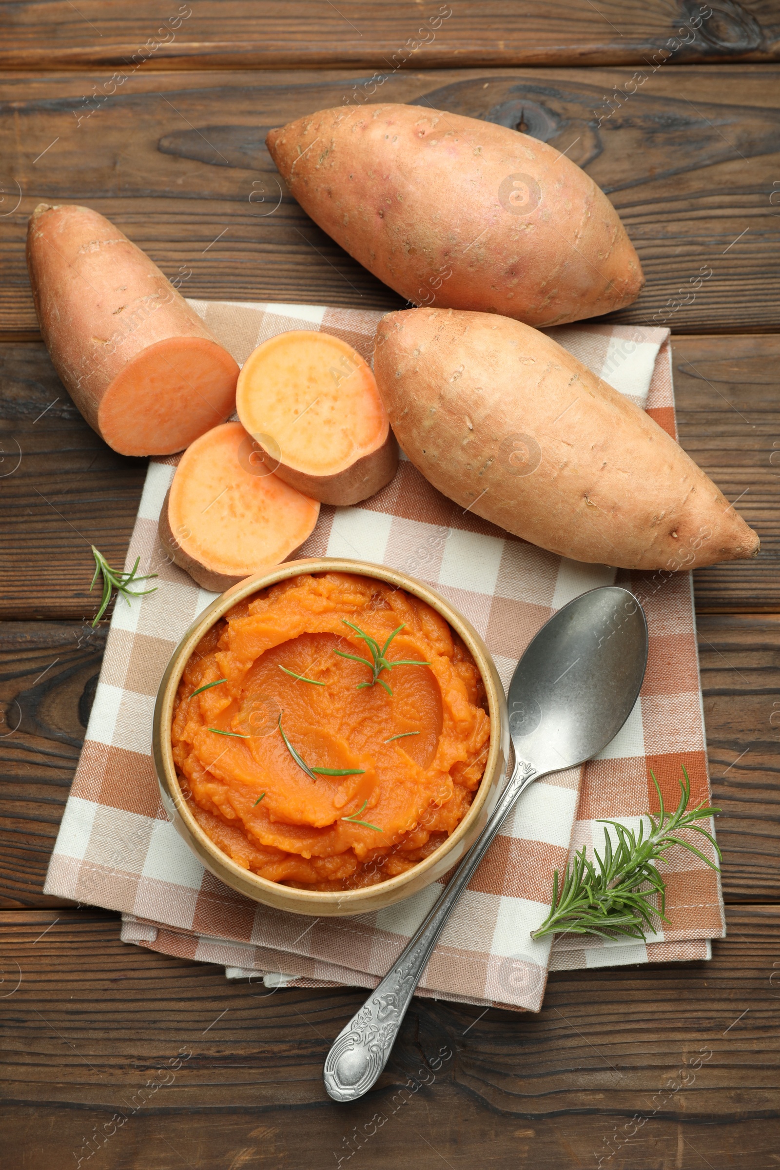 Photo of Tasty mashed sweet potato with rosemary in bowl, fresh vegetables and spoon on wooden table, flat lay