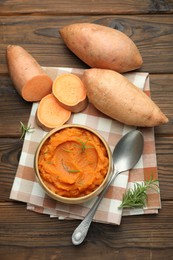 Photo of Tasty mashed sweet potato with rosemary in bowl, fresh vegetables and spoon on wooden table, flat lay