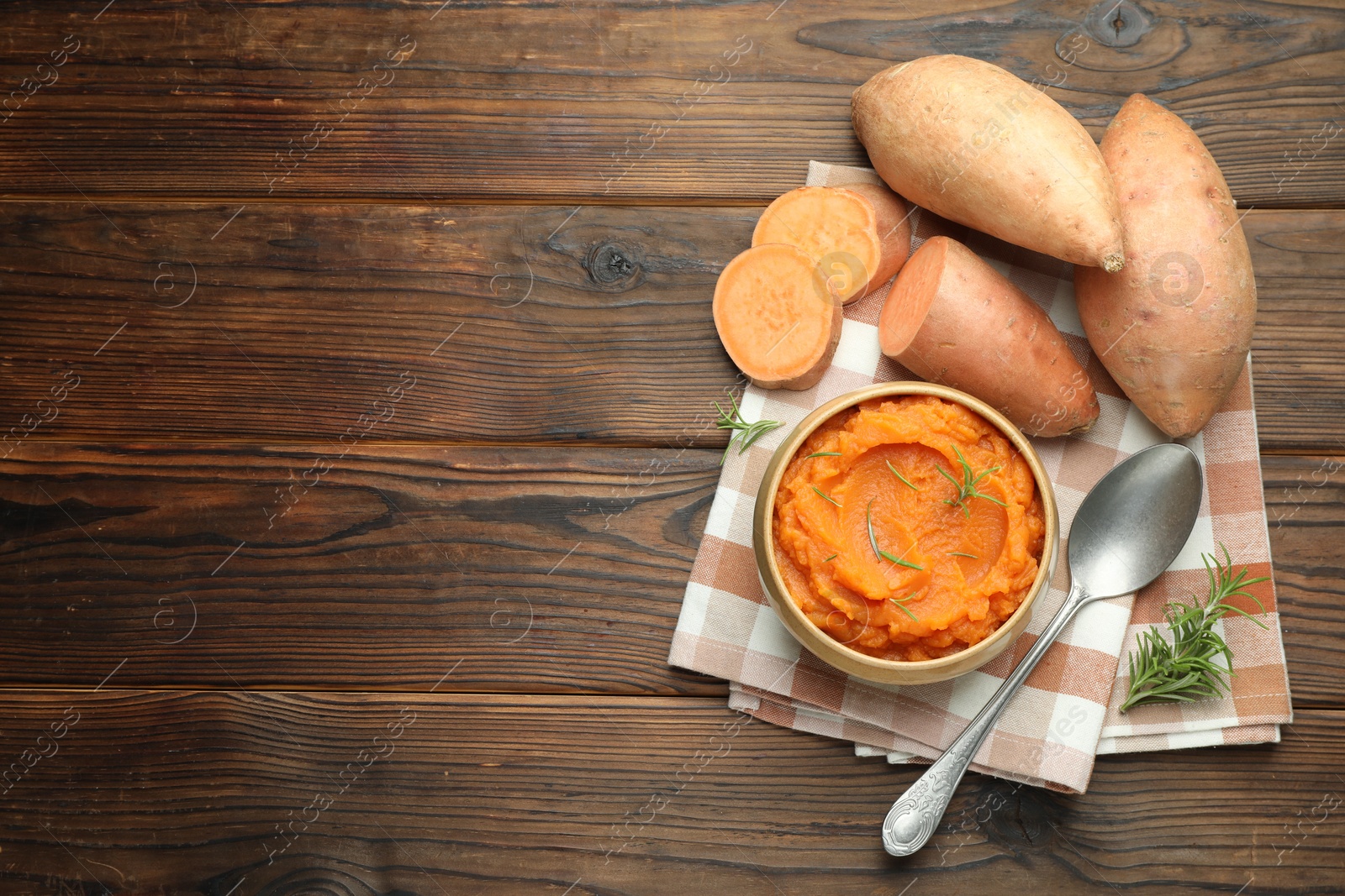 Photo of Tasty mashed sweet potato with rosemary in bowl, fresh vegetables and spoon on wooden table, flat lay. Space for text