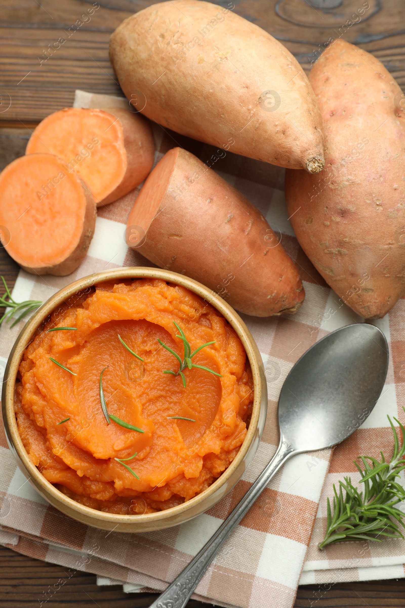 Photo of Tasty mashed sweet potato with rosemary in bowl, fresh vegetables and spoon on wooden table, flat lay