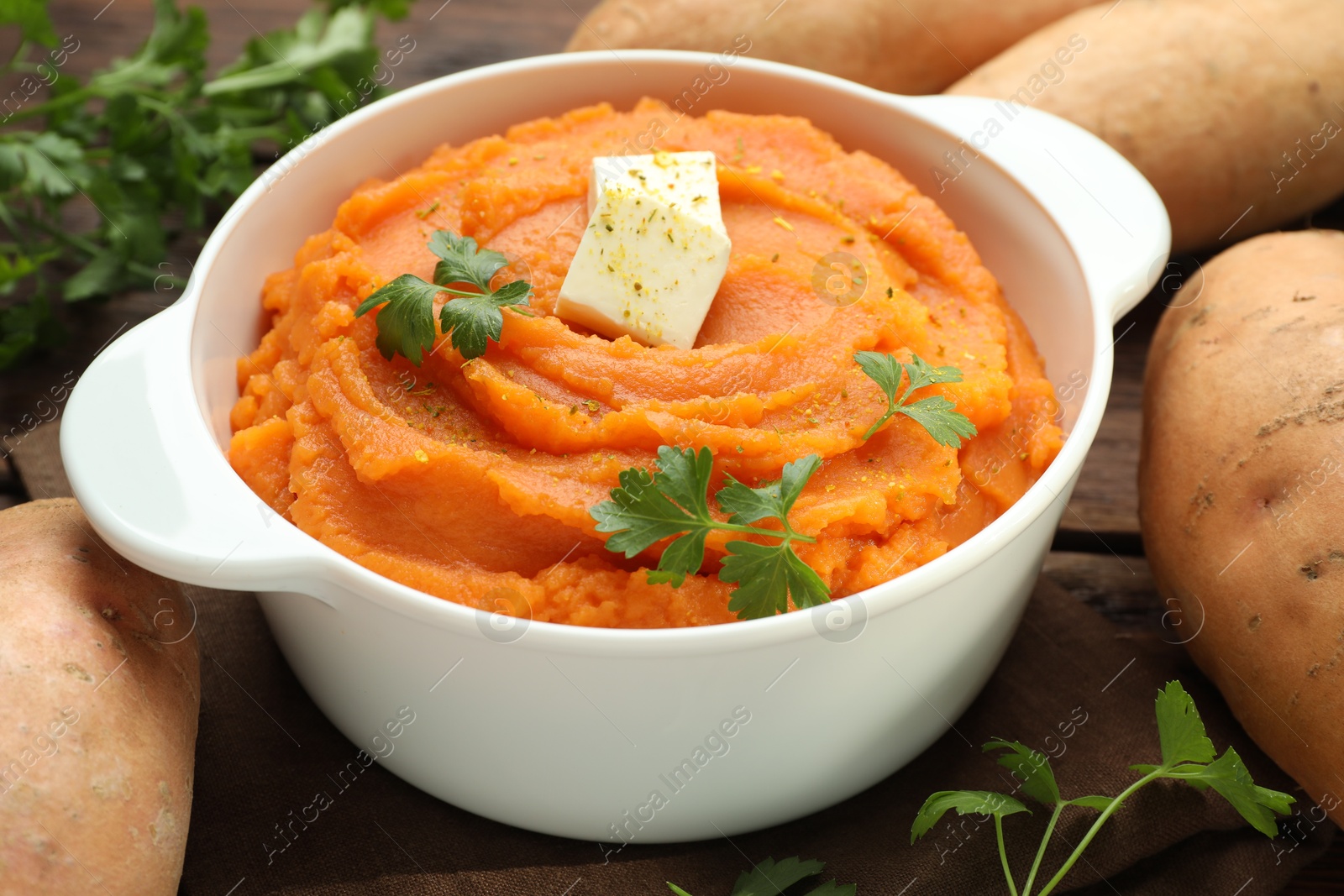 Photo of Tasty mashed sweet potato with butter in bowl, fresh vegetables and parsley on table, closeup