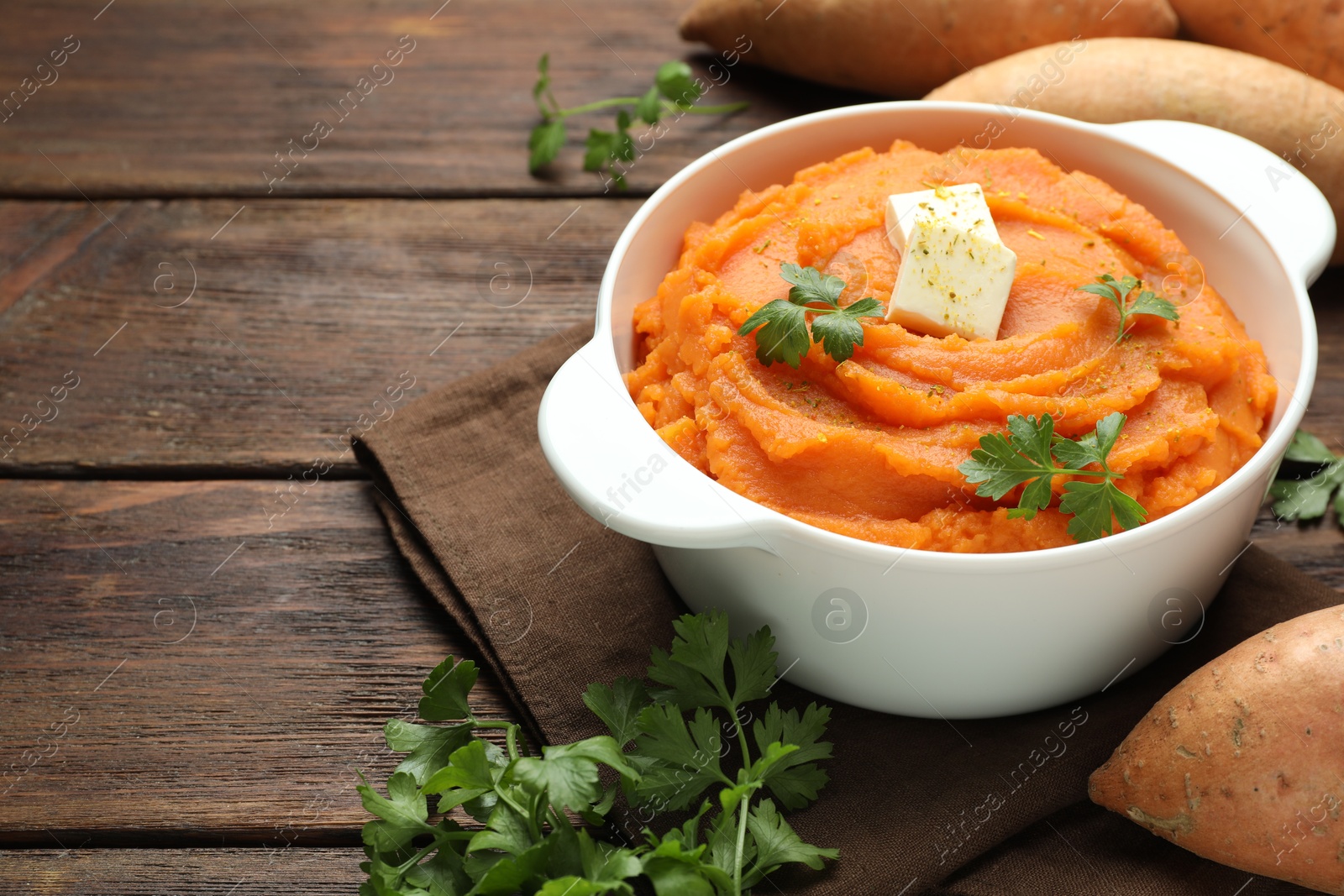 Photo of Tasty mashed sweet potato with butter in bowl, fresh vegetables and parsley on wooden table, closeup. Space for text