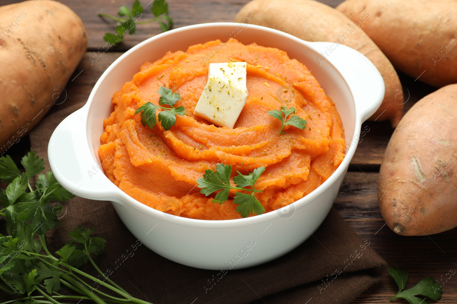 Photo of Tasty mashed sweet potato with butter in bowl, fresh vegetables and parsley on wooden table, closeup