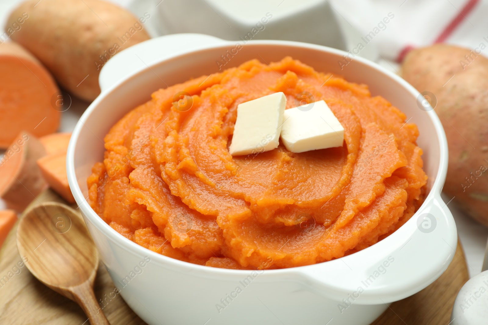 Photo of Tasty mashed sweet potato with butter in bowl, spoon and fresh vegetables on table, closeup