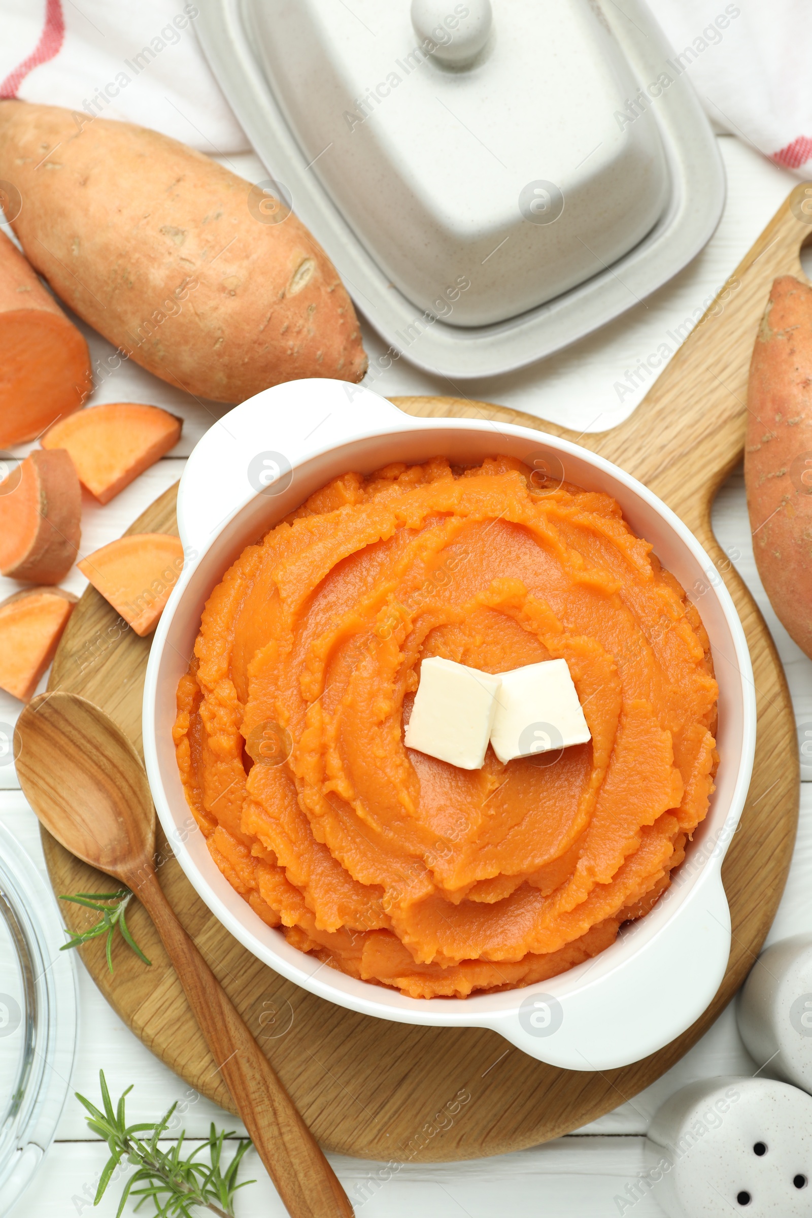 Photo of Tasty mashed sweet potato with butter in bowl, spoon, cut vegetables and spices on white wooden table, flat lay