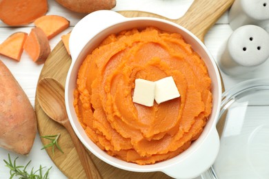 Photo of Tasty mashed sweet potato with butter in bowl, spoon, cut vegetables and spices on white wooden table, flat lay