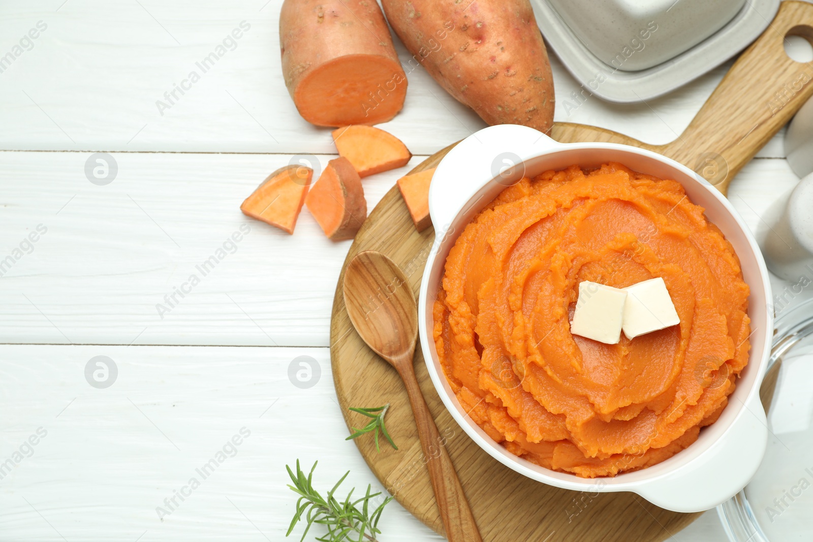 Photo of Tasty mashed sweet potato with butter in bowl, spoon, fresh vegetables and rosemary on white wooden table, flat lay. Space for text