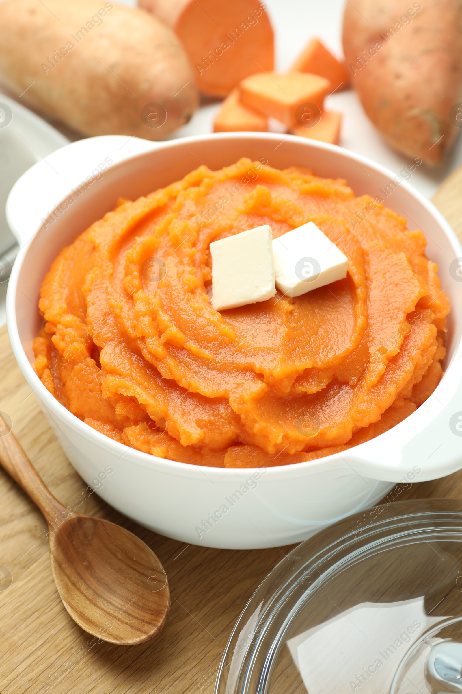 Photo of Tasty mashed sweet potato with butter in bowl, spoon and fresh vegetables on table, closeup