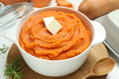 Photo of Tasty mashed sweet potato with butter in bowl, spoon, fresh vegetables and rosemary on table, closeup