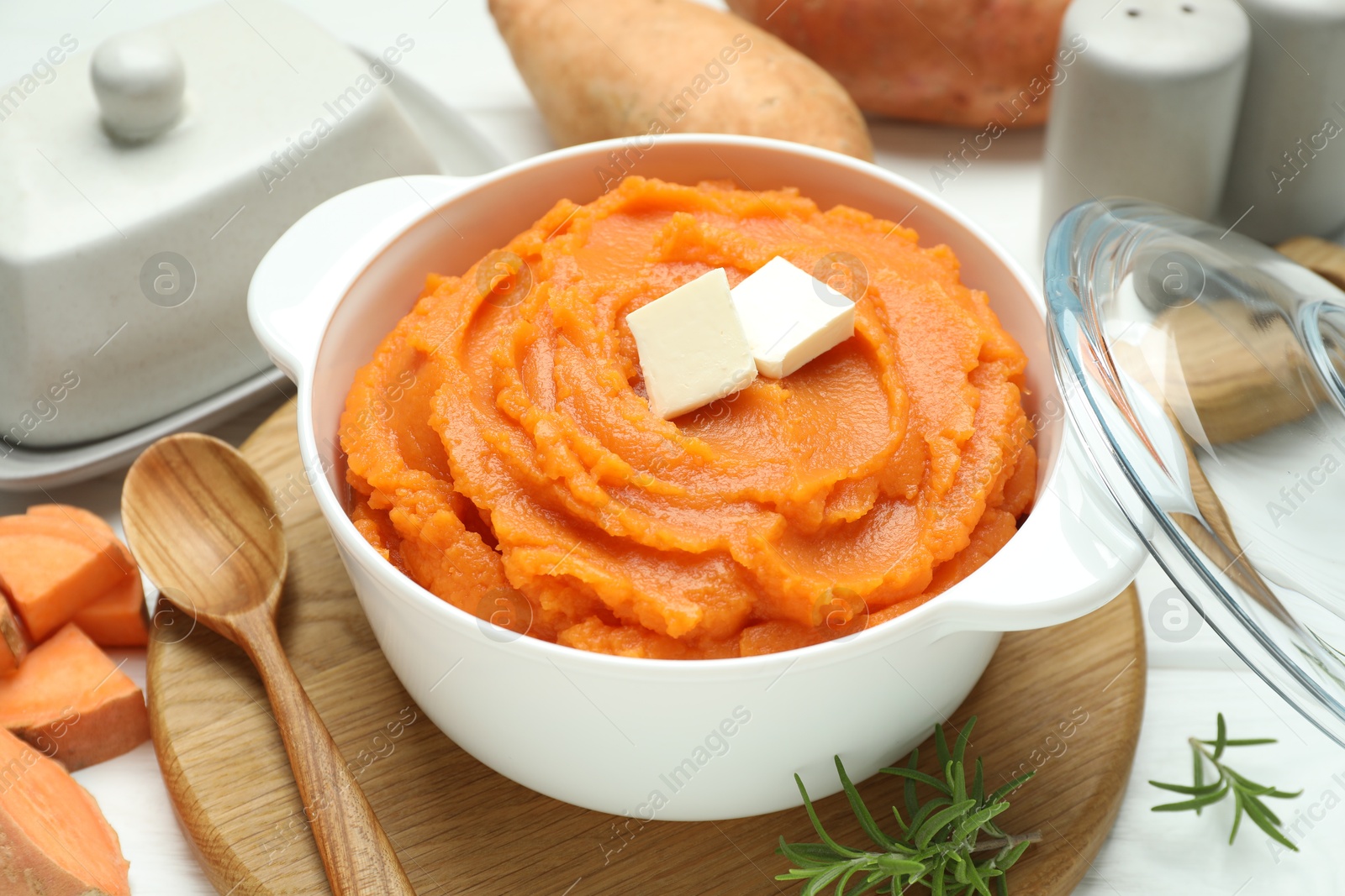 Photo of Tasty mashed sweet potato with butter in bowl, spoon, fresh vegetables and rosemary on table, closeup