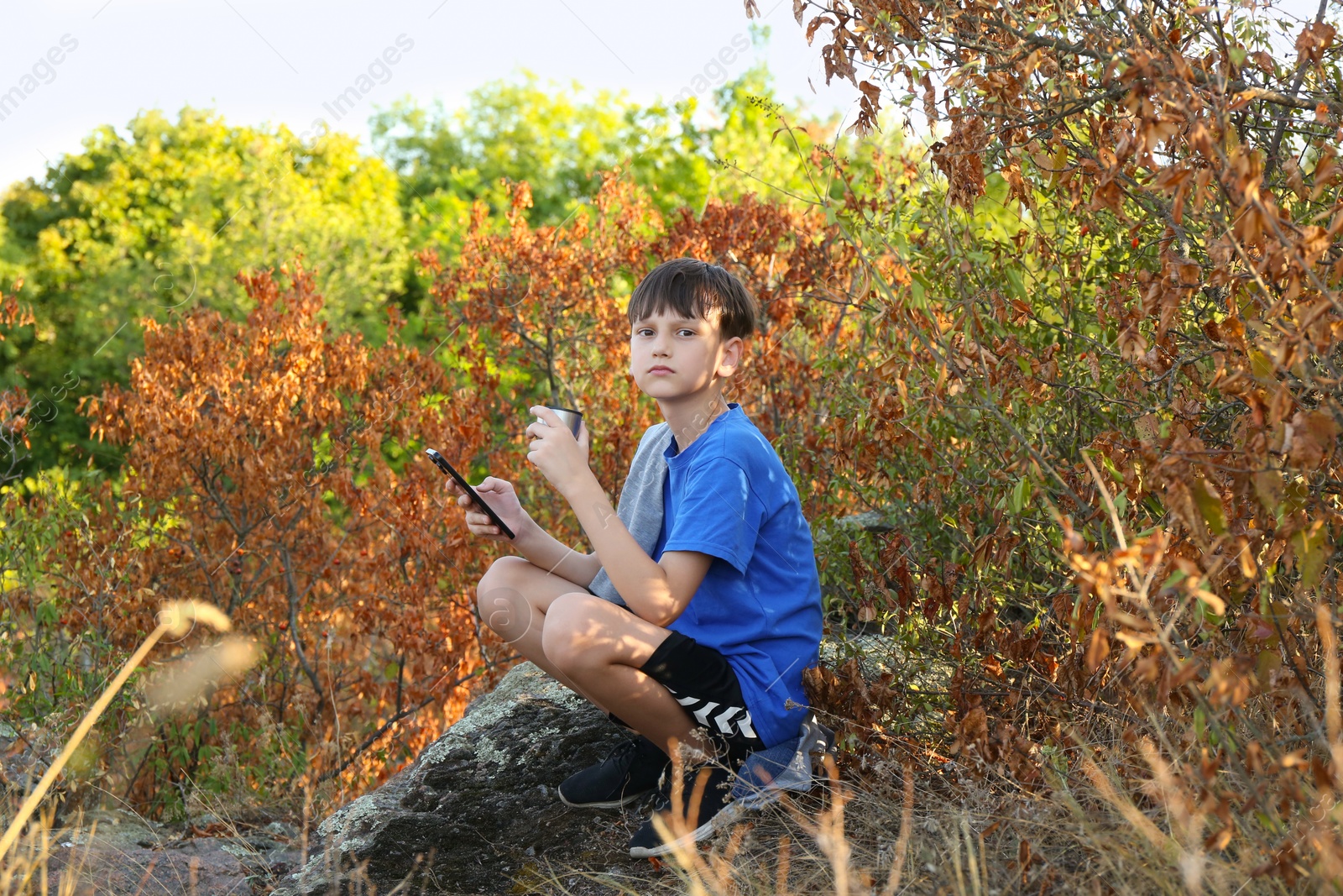 Photo of Little boy with smartphone drinking tea among trees outdoors