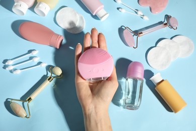 Woman holding facial cleansing brush near other body care products on light blue background, top view