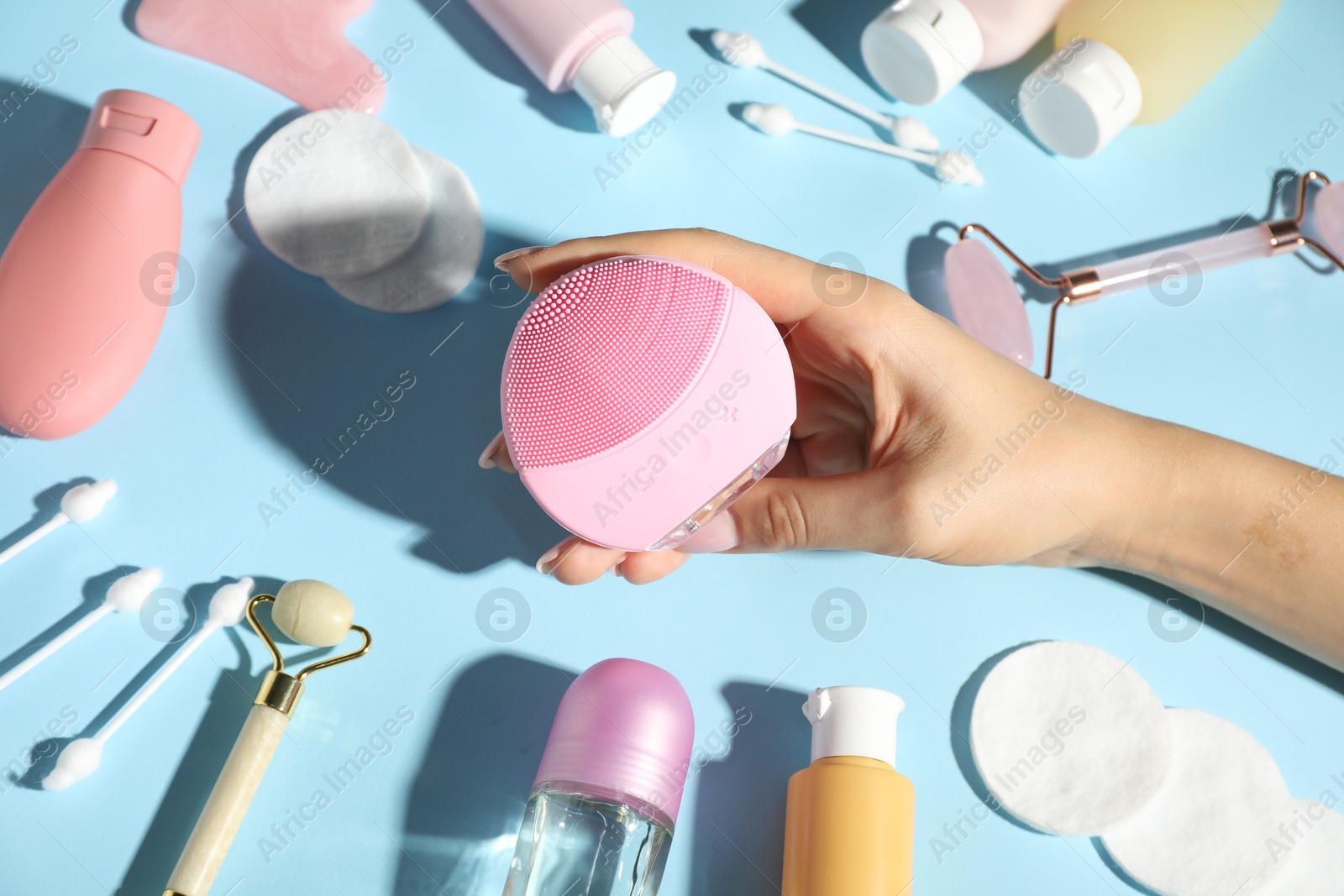 Photo of Woman holding facial cleansing brush near other body care products on light blue background, above view