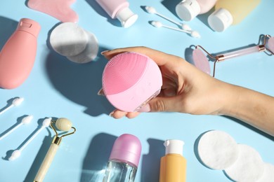 Woman holding facial cleansing brush near other body care products on light blue background, above view