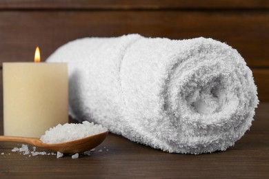 Photo of Spa composition. Towel, burning candle and spoon with sea salt on wooden table, closeup